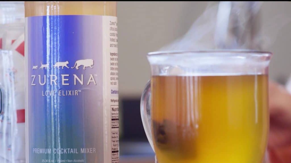 A picture of a bottle of Zurena Love Elixir alongside a hot glass of amber colored drink